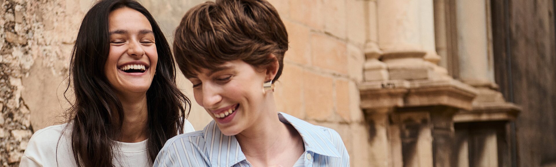 myGabor | Zwei lachende Frauen stehen leicht versetzt hintereinander vor einer alten Steinmauer, Nahaufnahme der Oberkörper und Gesichter | © Gabor Shoes GmbH, Rosenheim