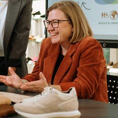 Une femme en blazer orange est assise à une table et gesticule avec ses mains. Sur la table se trouve une basket claire. À l’arrière-plan, on voit un écran avec une présentation. | © Gabor Shoes GmbH, Allemagne