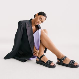 Une femme est assise dans une pièce lumineuse, porte une robe lilas et un blazer noir ainsi que des sandales noires à brides et détails décoratifs. | © Gabor Shoes GmbH, Allemagne