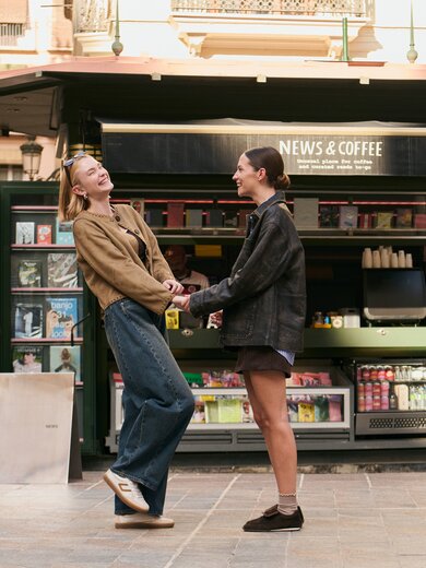 Deux femmes se tiennent devant un kiosque ouvert portant l’enseigne "News & Coffee", elles se tiennent par la main et se sourient. À l’arrière-plan, on voit des magazines et des boissons. | © Gabor Shoes GmbH, Allemagne