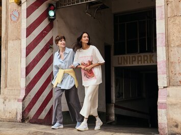 Deux femmes sortent bras dessus bras dessous d’un parking souterrain sur le trottoir, à côté d’un mur à rayures rouges et blanches et d’un feu vert. Elles portent toutes les deux des vêtements décontractés et des baskets claires. | © Gabor Shoes GmbH, Allemagne