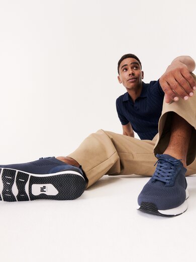 Un homme est assis de manière détendue sur le sol, porte un pantalon beige, un haut bleu et des baskets bleu foncé. Les chaussures sont visibles au premier plan de l’image. | © Gabor Shoes GmbH, Allemagne