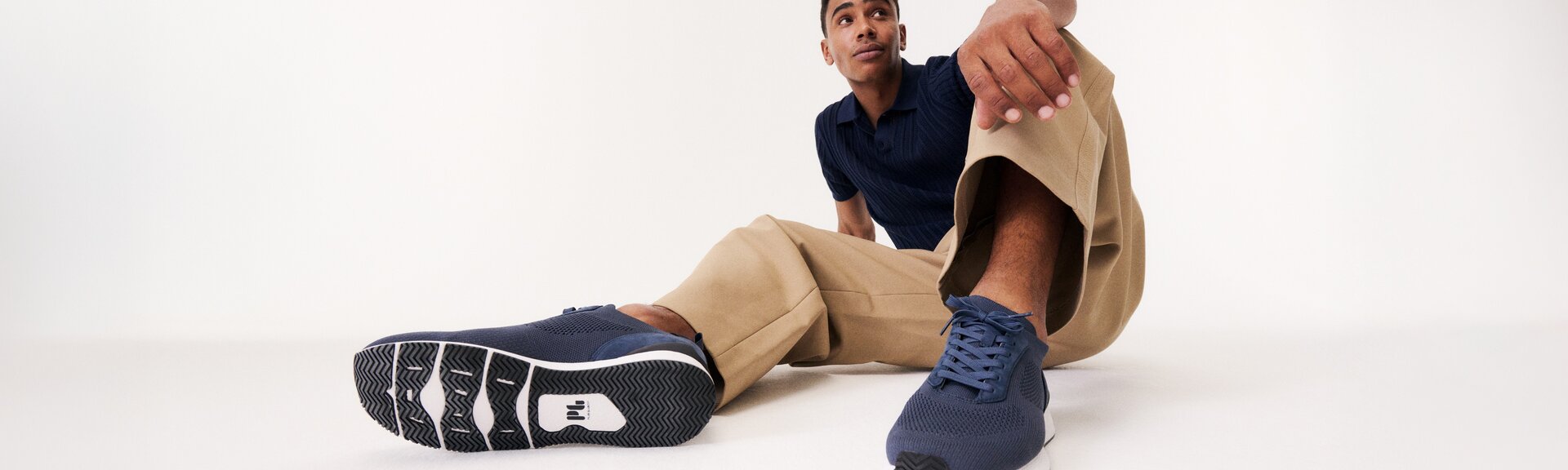 Un homme est assis de manière détendue sur le sol, porte un pantalon beige, un haut bleu et des baskets bleu foncé. Les chaussures sont visibles au premier plan de l’image. | © Gabor Shoes GmbH, Allemagne