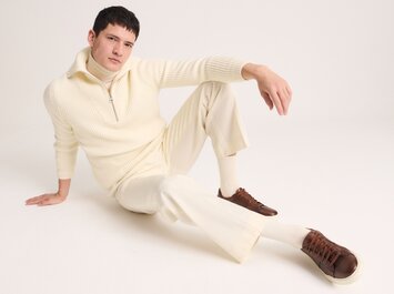 Un homme est assis détendu par terre et porte un pull en maille crème à fermeture éclair, un pantalon crème assorti et des sneakers marron. L’arrière-plan est sobre et clair. | © Gabor Shoes GmbH, Allemagne