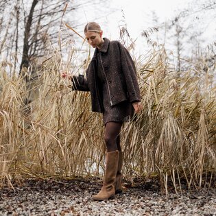 Une femme se tient dans la nature automnale sur un chemin de gravier, porte des bottes marron, une veste marron et une jupe courte, et s’appuie contre de hautes roseaux secs. | © Gabor Shoes GmbH, Allemagne