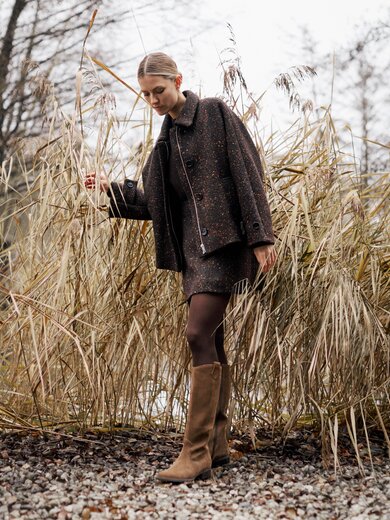 Une femme se tient dans la nature automnale sur un chemin de gravier, porte des bottes marron, une veste marron et une jupe courte, et s’appuie contre de hautes roseaux secs. | © Gabor Shoes GmbH, Allemagne