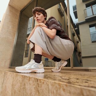 Une femme est accroupie sur une marche en pierre devant un bâtiment moderne et porte des baskets claires, des chaussettes foncées, une jupe ample et un pull à manches courtes. | © Gabor Shoes GmbH, Allemagne