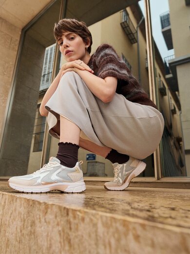 Une femme est accroupie sur une marche en pierre devant un bâtiment moderne et porte des baskets claires, des chaussettes foncées, une jupe ample et un pull à manches courtes. | © Gabor Shoes GmbH, Allemagne