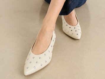 Une femme est assise par terre et porte un pantalon bleu foncé ainsi que des escarpins beiges pointus avec des clous argentés. | © Gabor Shoes GmbH, Allemagne