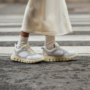 Deux jambes en chaussettes de sport blanches et baskets crème se tiennent sur un passage piéton. La femme porte une longue jupe claire. | © Gabor Shoes GmbH, Allemagne