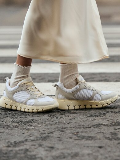 Deux jambes en chaussettes de sport blanches et baskets crème se tiennent sur un passage piéton. La femme porte une longue jupe claire. | © Gabor Shoes GmbH, Allemagne
