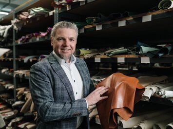 Markus Kramer, directeur des achats chez Gabor, tient un grand morceau de cuir brun à la main et se tient devant une étagère avec différents morceaux de cuir dans un entrepôt. | © Gabor Shoes GmbH, Allemagne