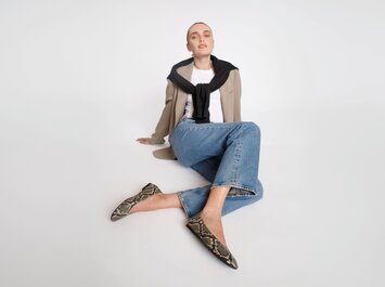 Une femme est assise par terre, porte un jean bleu, un t-shirt blanc, un blazer beige, un pull noir sur les épaules et des chaussures plates à motif serpent. L’arrière-plan est blanc et épuré. | © Gabor Shoes GmbH, Allemagne