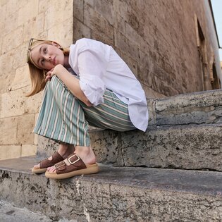 Une femme est assise sur un escalier en pierre devant un vieux mur. Elle porte un chemisier blanc, un pantalon ample rayé vert et beige, ainsi que des sandales marron à boucle dorée. Elle a glissé des lunettes de soleil dans ses cheveux. | © Gabor Shoes GmbH, Allemagne