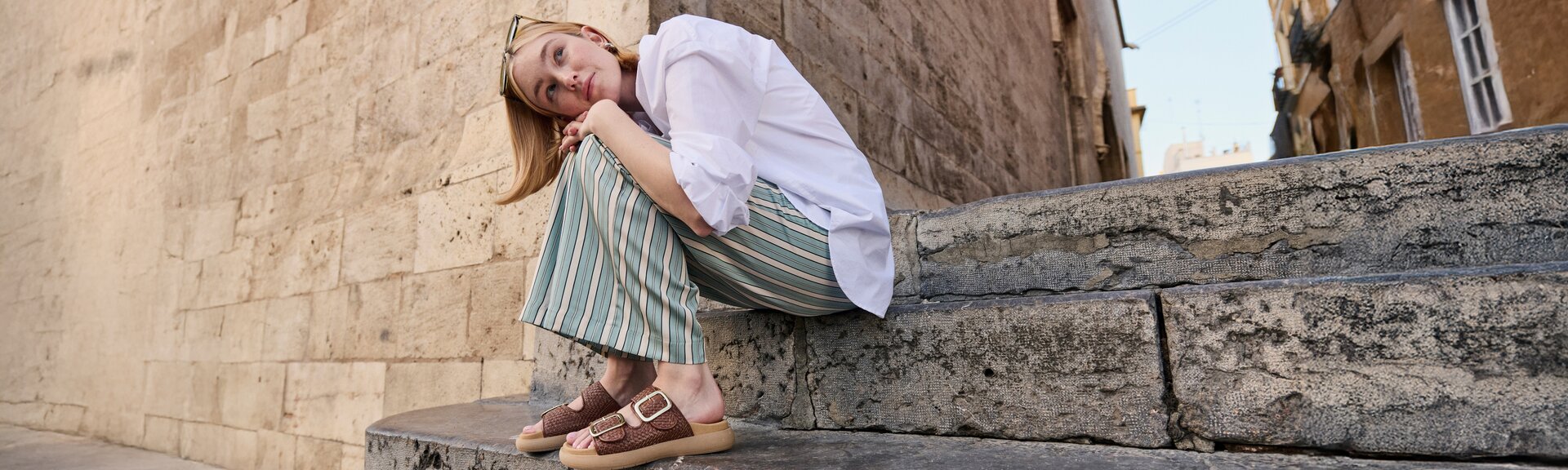 Une femme est assise sur un escalier en pierre devant un vieux mur. Elle porte un chemisier blanc, un pantalon ample rayé vert et beige, ainsi que des sandales marron à boucle dorée. Elle a glissé des lunettes de soleil dans ses cheveux. | © Gabor Shoes GmbH, Allemagne