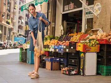Une femme en robe en jean et sandales se tient devant un stand de fruits et légumes sur le trottoir et tient un filet d’oranges. À l’arrière-plan, on aperçoit des caisses de fruits colorées et une rue avec des bâtiments. | © Gabor Shoes GmbH, Allemagne
