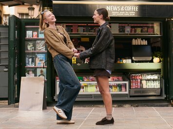 Deux femmes se tiennent devant un kiosque ouvert portant l’enseigne "News & Coffee", elles se tiennent par la main et se sourient. À l’arrière-plan, on voit des magazines et des boissons. | © Gabor Shoes GmbH, Allemagne