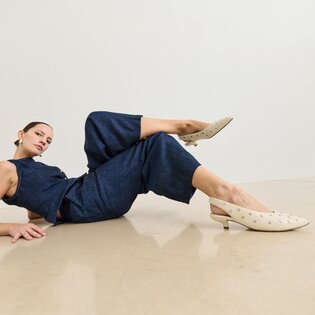 Une femme porte une combinaison bleu foncé et est allongée de manière détendue sur le sol. Les escarpins beiges avec de petits clous argentés et un talon bas sont au centre de l’image. | © Gabor Shoes GmbH, Allemagne