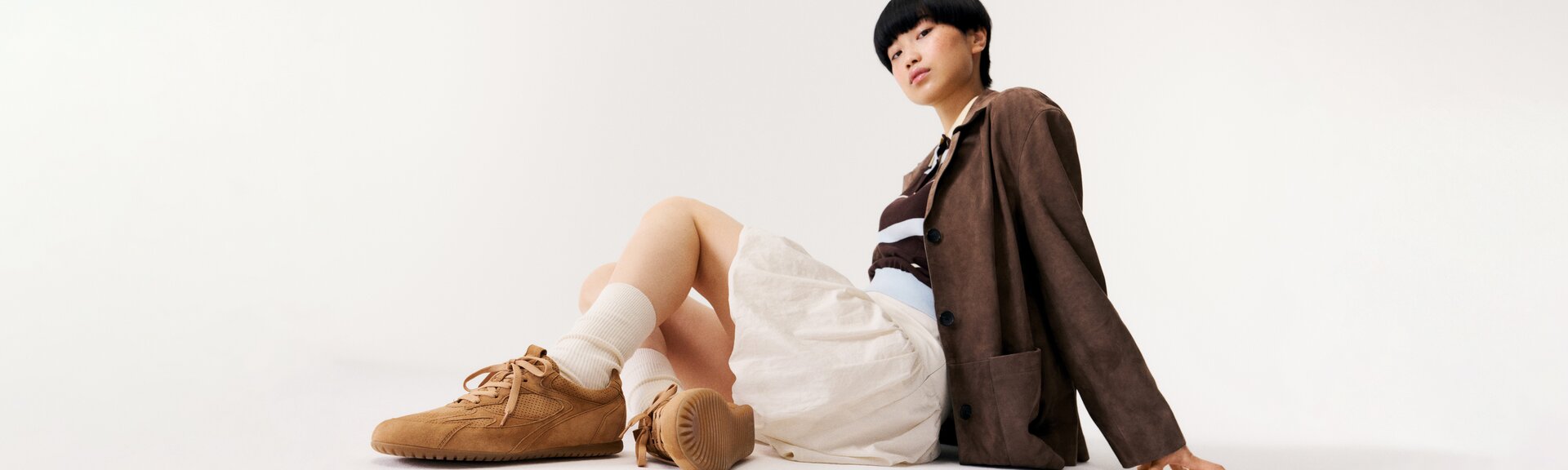 Une femme est assise par terre devant un fond blanc et porte des baskets marron, des chaussettes blanches, une jupe beige, un haut rayé et un blazer marron. | © Gabor Shoes GmbH, Allemagne
