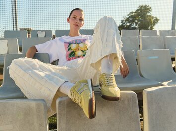 Une femme est assise détendue sur des gradins de stade, porte un pantalon clair à texture, un t-shirt blanc avec un motif de citron et des baskets jaunes. | © Gabor Shoes GmbH, Allemagne
