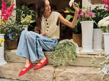 Une femme est assise sur des marches en pierre devant un fleuriste, entourée de fleurs colorées et de vases blancs. Elle porte un gilet en tricot clair, un jean ample et des chaussures rouges voyantes. Elle tient une tige de fleur à la main. | © Gabor Shoes GmbH, Allemagne