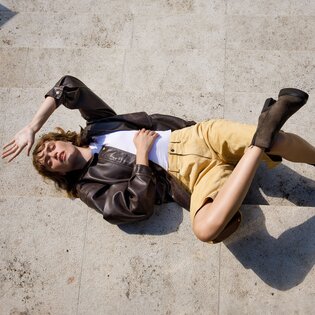 Une femme est allongée sur des marches en pierre. Elle porte une veste en cuir marron, un haut blanc, un short beige et des bottines marron. | © Gabor Shoes GmbH, Allemagne