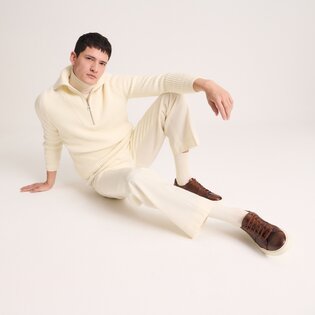Un homme est assis détendu par terre et porte un pull en maille crème à fermeture éclair, un pantalon crème assorti et des sneakers marron. L’arrière-plan est sobre et clair. | © Gabor Shoes GmbH, Allemagne