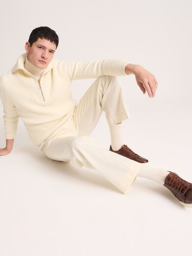 Un homme est assis détendu par terre et porte un pull en maille crème à fermeture éclair, un pantalon crème assorti et des sneakers marron. L’arrière-plan est sobre et clair. | © Gabor Shoes GmbH, Allemagne