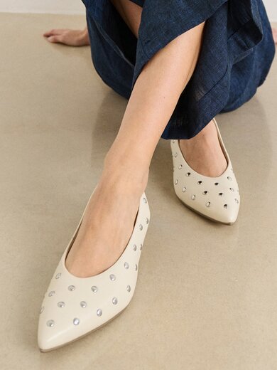 Une femme est assise par terre et porte un pantalon bleu foncé ainsi que des escarpins beiges pointus avec des clous argentés. | © Gabor Shoes GmbH, Allemagne