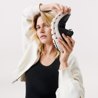 Une femme tient une basket noire flexible à semelle blanche près de sa tête. Elle porte une veste blanche et un haut noir devant un fond clair. | © Gabor Shoes GmbH, Allemagne