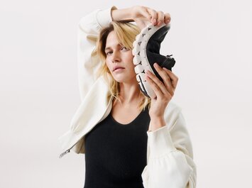Une femme tient une basket noire flexible à semelle blanche près de sa tête. Elle porte une veste blanche et un haut noir devant un fond clair. | © Gabor Shoes GmbH, Allemagne