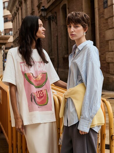 Deux femmes se tiennent côte à côte sur une rue mouillée devant un vieux bâtiment ; l’une porte un t-shirt blanc avec un motif de pastèque et l’inscription « Tropical », l’autre une chemise bleu clair et un pull jaune noué autour des hanches. À l’arrière-plan, des vélos sont appuyés contre une barrière. | © Gabor Shoes GmbH, Allemagne