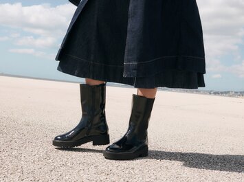 Une femme se tient dehors sur un sol clair, porte une jupe foncée et des bottes noires brillantes. Le ciel est bleu avec quelques nuages. | © Gabor Shoes GmbH, Allemagne
