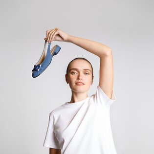 Une femme en t-shirt blanc tient une chaussure bleue avec petit talon et nœud en l’air, devant un fond clair. | © Gabor Shoes GmbH, Allemagne
