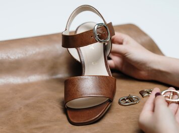 Une sandale marron avec une boucle argentée est présentée sur du cuir brun. Une main tient la sandale, plusieurs autres boucles sont posées à côté. | © Gabor Shoes GmbH, Allemagne