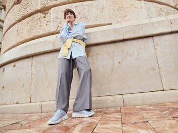Une femme se tient de façon décontractée devant un mur de pierre clair, porte un pantalon gris ample, des sneakers bleu clair, une longue chemise rayée bleue et a un pull jaune noué lâchement autour des hanches. Photographiée en contre-plongée. | © Gabor Shoes GmbH, Allemagne