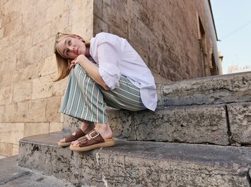 Une femme est assise sur un escalier en pierre devant un vieux mur. Elle porte un chemisier blanc, un pantalon ample rayé vert et beige, ainsi que des sandales marron à boucle dorée. Elle a glissé des lunettes de soleil dans ses cheveux. | © Gabor Shoes GmbH, Allemagne