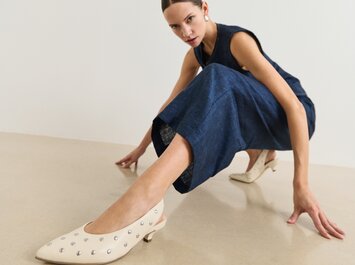 Une femme en combinaison bleu foncé sans manches présente des chaussures couleur crème avec de petits clous argentés et un talon bas sur un sol clair devant un fond blanc. | © Gabor Shoes GmbH, Allemagne