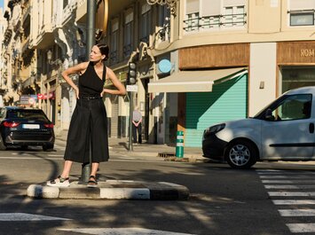 Une femme se tient sur un îlot central à un carrefour, porte un haut noir sans manches, un pantalon culotte ample noir et des sandales. À l’arrière-plan, on voit des voitures, des maisons et un passage piéton. | © Gabor Shoes GmbH, Allemagne