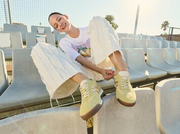 Une femme portant un pantalon blanc évasé et des baskets jaunes est assise au soleil sur des sièges de stade vides et enfile sa chaussure gauche. | © Gabor Shoes GmbH, Allemagne