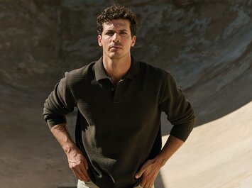 Un homme se tient les mains sur les hanches devant un fond rond en béton, et porte un pull en maille vert foncé avec col et un pantalon beige. | © Gabor Shoes GmbH, Allemagne