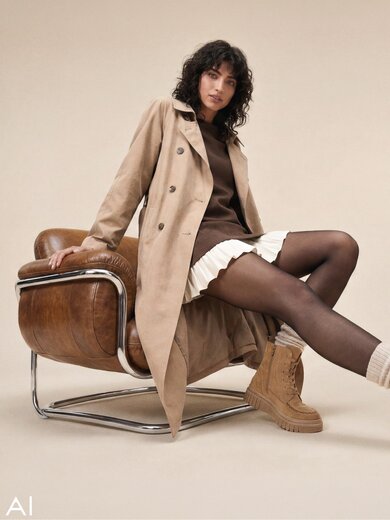 En AI-genererad bild som visar en kvinna som sitter i en läderfåtölj framför en beige bakgrund. Hon bär en ljusbrun trenchcoat över en mörkbrun topp och en vit plisserad kjol och har beige snörstövlar från Gabor på fötterna. | © Gabor Shoes GmbH, Tyskland
