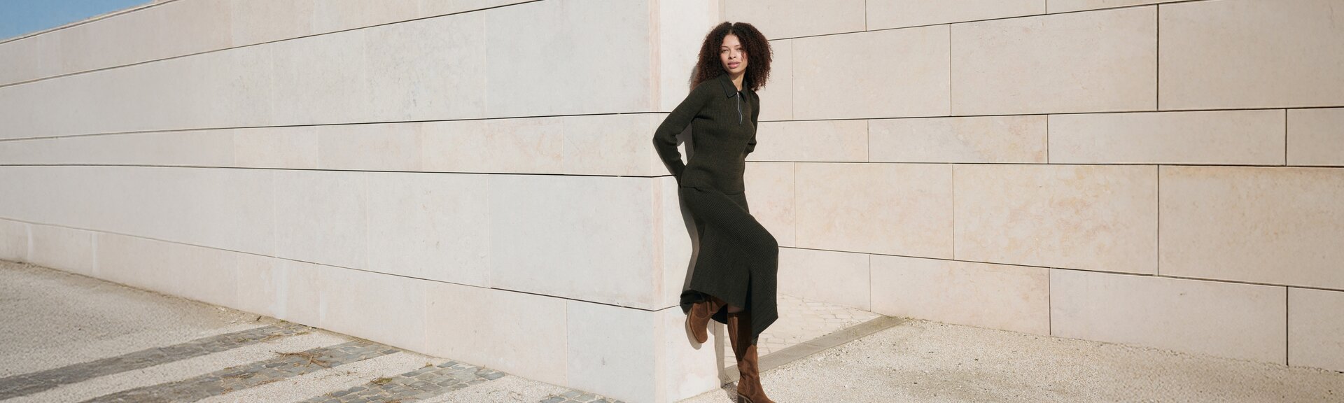 Une femme se tient détendue devant un mur de pierre clair, porte une longue robe sombre et des bottes marron. L’image a été prise en extérieur à la lumière du jour. | © Gabor Shoes GmbH, Allemagne