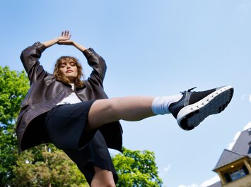 Une femme se tient à l’extérieur, porte une veste foncée, une jupe noire et des baskets noires, une jambe est levée en l’air. | © Gabor Shoes GmbH, Allemagne