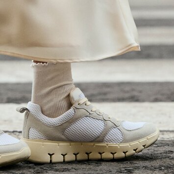 Deux jambes en chaussettes de sport blanches et baskets crème se tiennent sur un passage piéton. La femme porte une longue jupe claire. | © Gabor Shoes GmbH, Allemagne