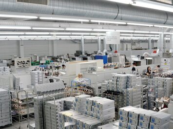 Grand hall de production avec étagères, cartons, machines et plusieurs personnes au travail.