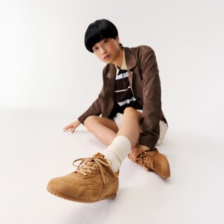 Une femme est assise par terre dans une pièce blanche et porte des baskets marron, des chaussettes blanches, une jupe beige, un haut rayé et un blazer marron. | © Gabor Shoes GmbH, Allemagne