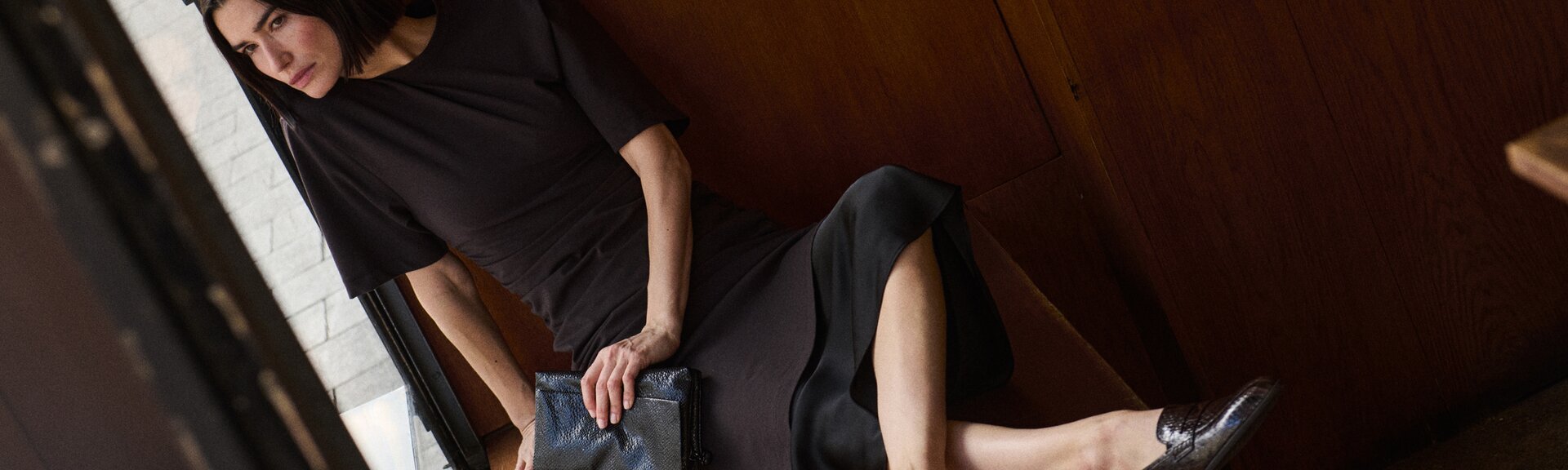 Een vrouw staat ontspannen bij een lichte stenen muur, draagt een donker lang kleed en bruine laarzen. De foto is buiten bij daglicht genomen. | © Gabor Shoes GmbH, Duitsland