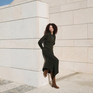 Une femme aux cheveux bouclés s’appuie contre un mur de pierre clair, porte une longue robe noire et des bottes marron. La scène est ensoleillée et calme. | © Gabor Shoes GmbH, Allemagne