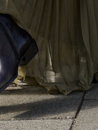 Una donna indossa stivaletti marrone scuro da donna con punta affusolata e tacco basso abbinati a una lunga gonna fluida verde oliva; è in piedi su un marciapiede lastricato davanti a dei gradini in pietra. | © Gabor Shoes GmbH, Germania